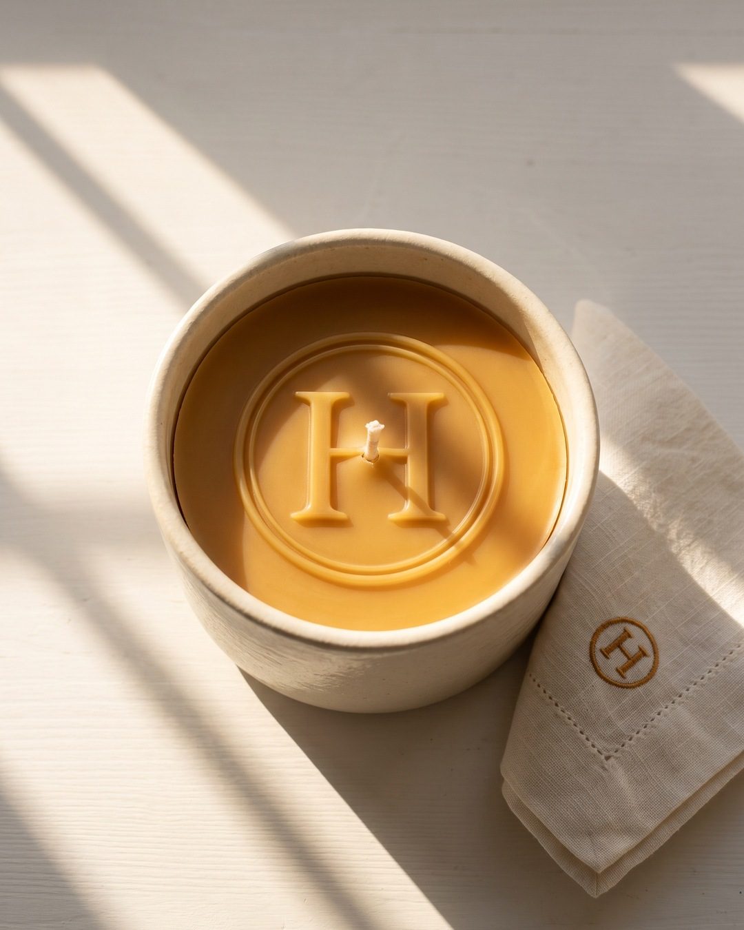 A honey-coloured candle with a large H monogram pressed into its wax, beside a linen napkin stitched with the same mark