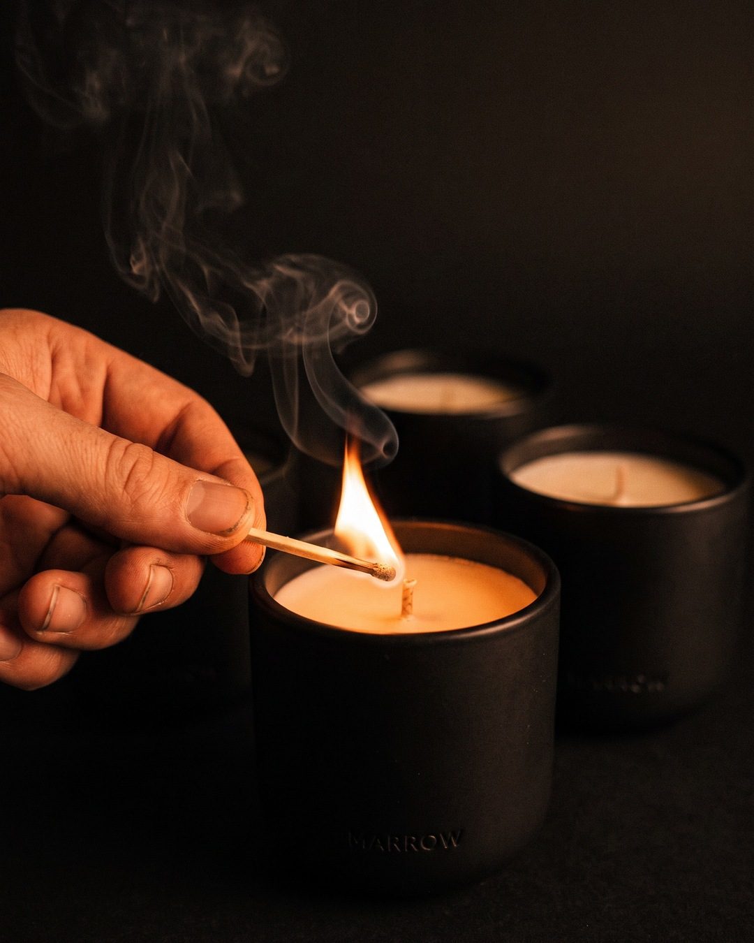 A hand striking a match to light a black ceramic candle in a dark room, smoke curling up