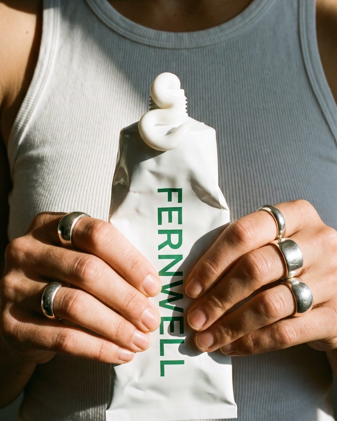 Hands with chunky silver rings squeezing a white Fernwell tube so a curl of cream rises from the top