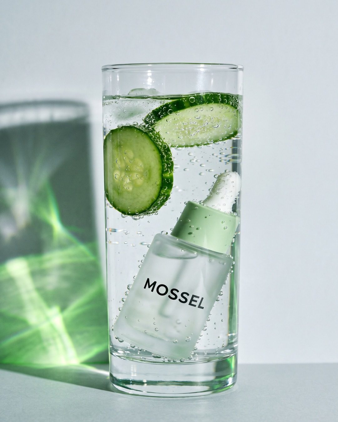 A frosted Mossel serum bottle in a tall glass of sparkling water with cucumber slices, green light rippling on the wall