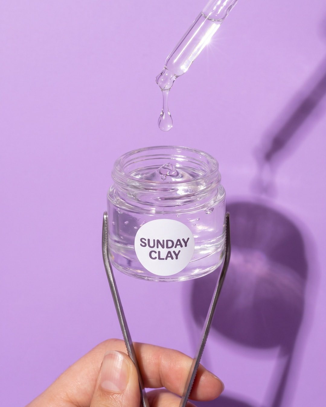 A clear serum drop falling from a pipette into a Sunday Clay gel jar held in tweezers on a lilac background