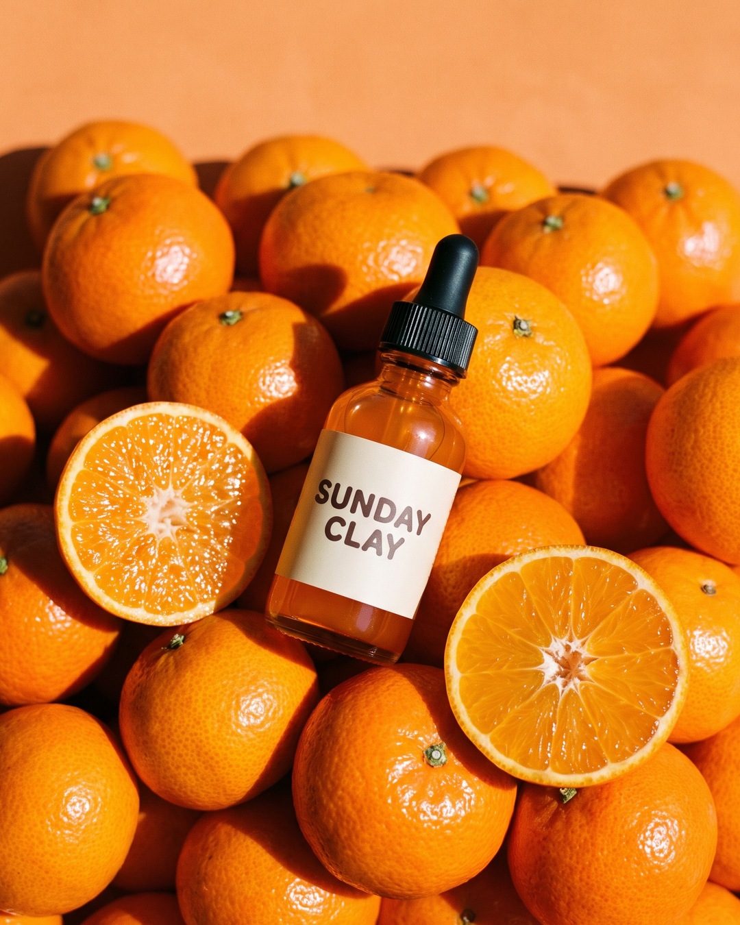 An orange glass Sunday Clay serum bottle resting on a heap of clementines, two of them cut in half