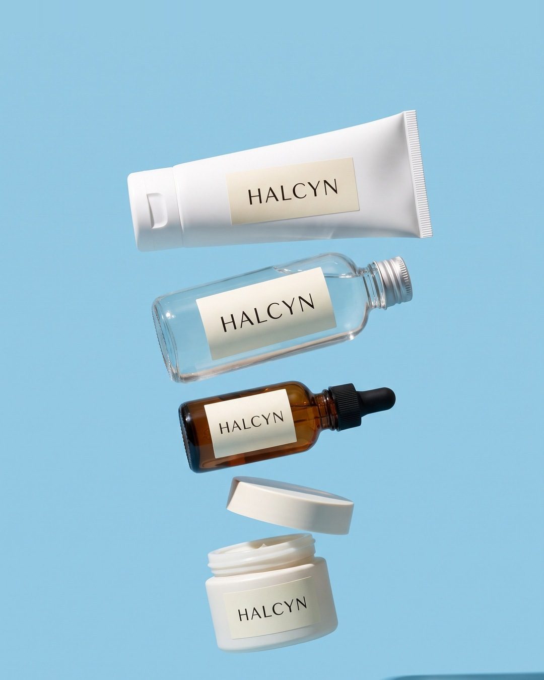 Four Halcyn skincare products floating in a vertical stack on sky blue: a tube, a toner, a serum dropper and a jar