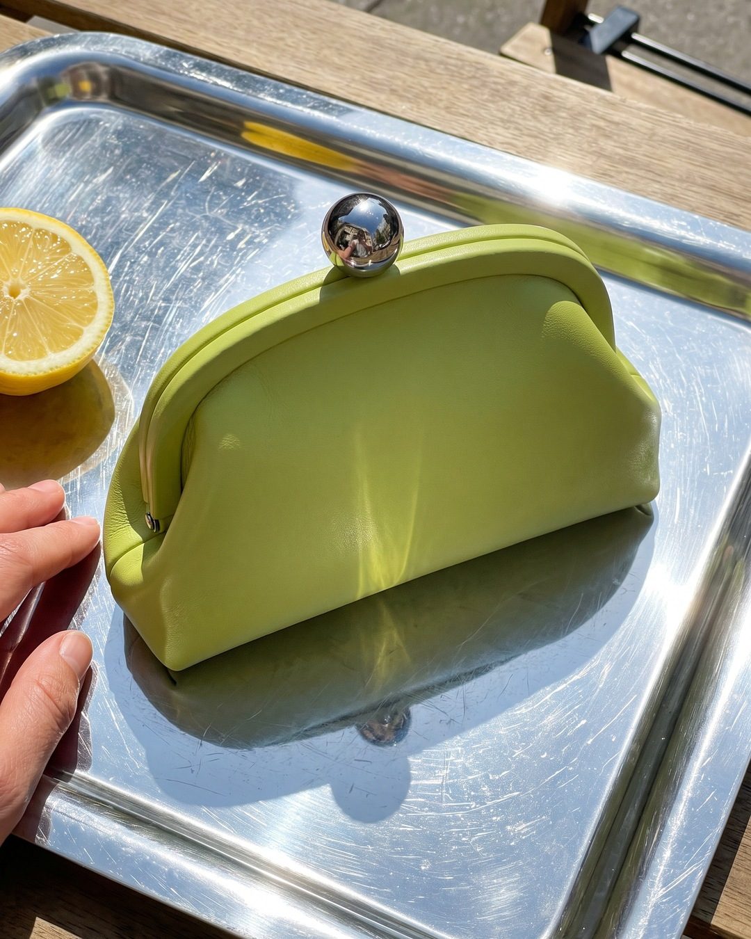 A lime-green frame clutch with a chrome ball clasp on a shiny silver tray in the sun, beside half a lemon