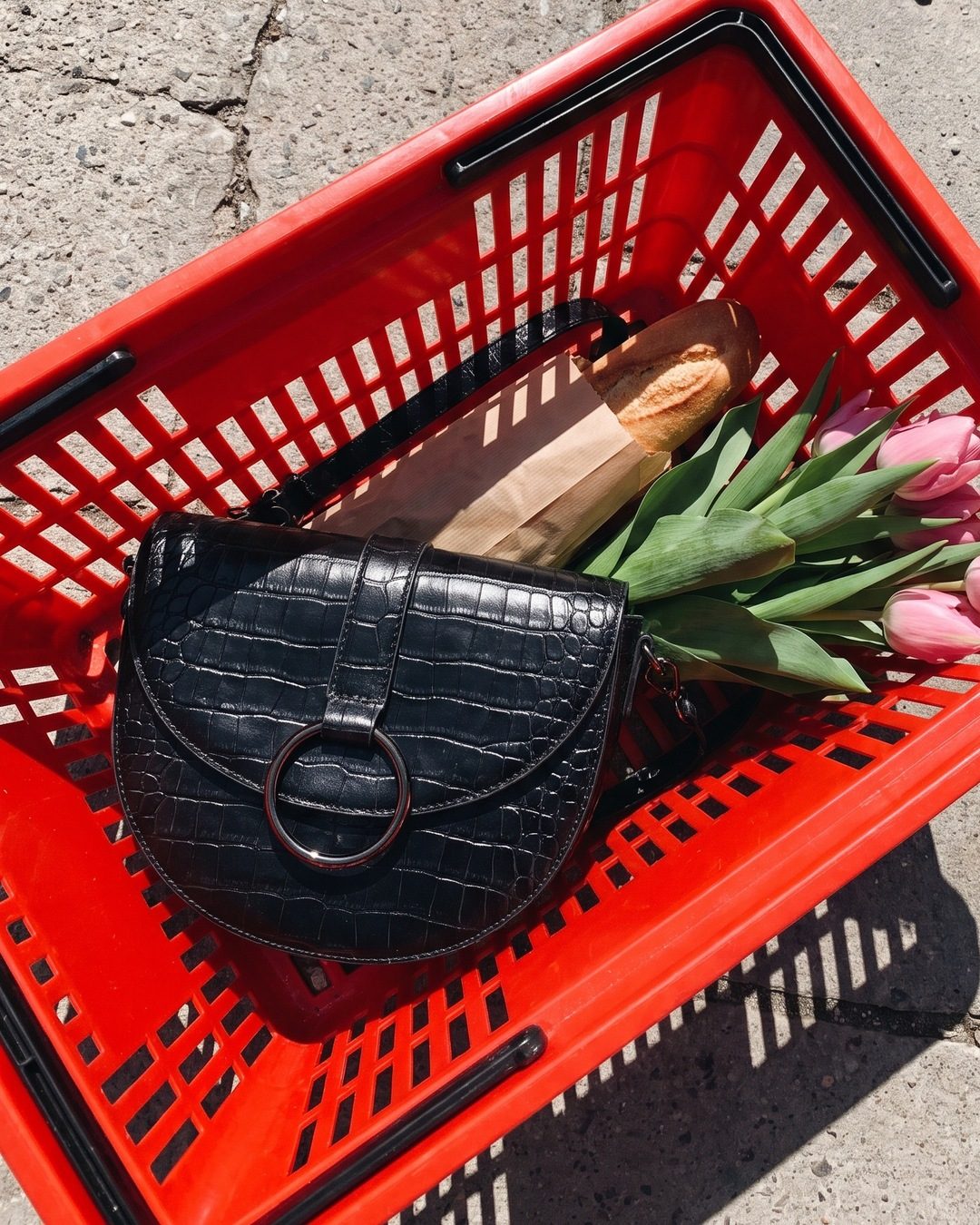 A black croc-embossed half-moon bag in a red shopping basket with a baguette and pink tulips