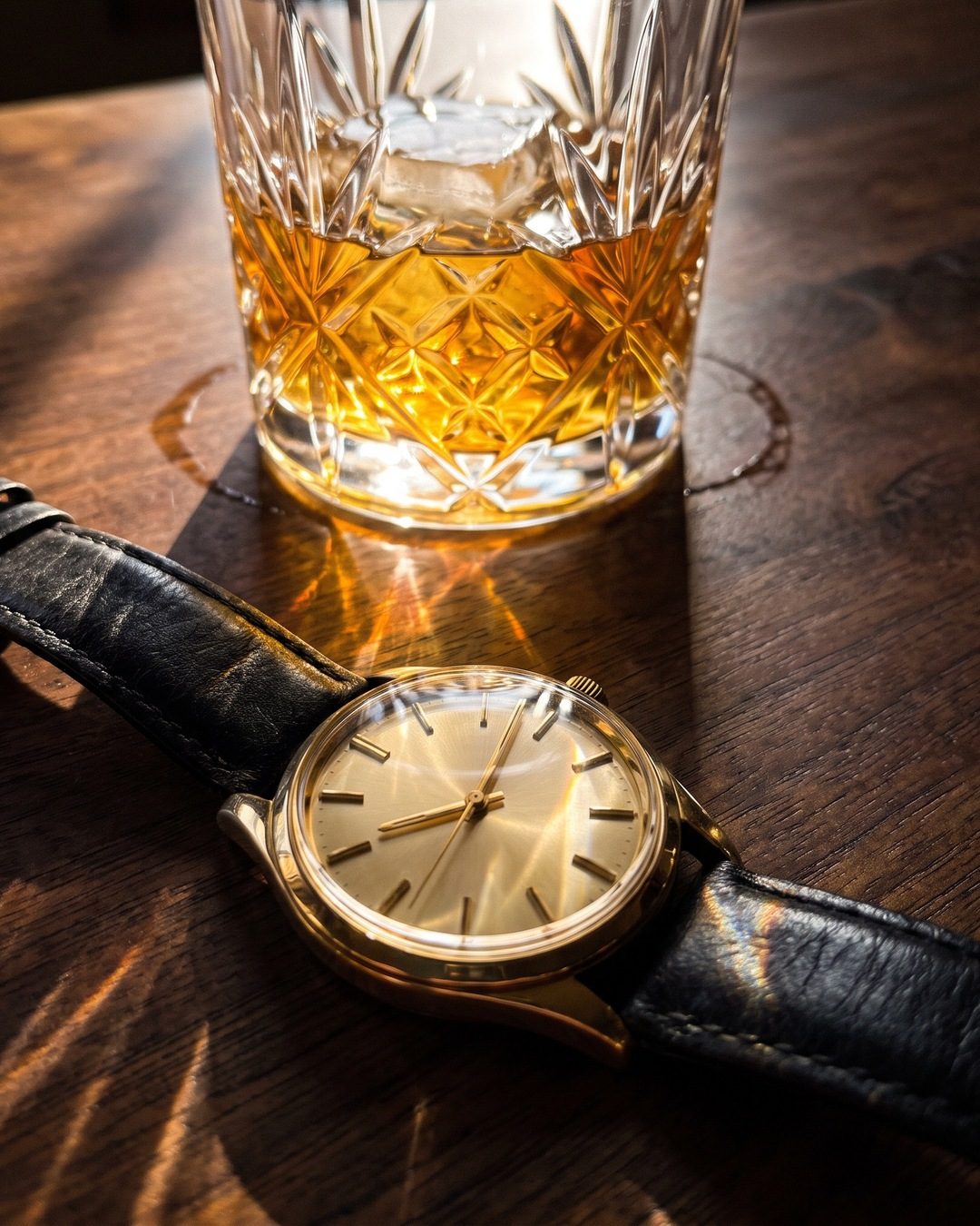 A gold watch on a walnut bar top, with sun through a crystal tumbler of whisky throwing amber light across it