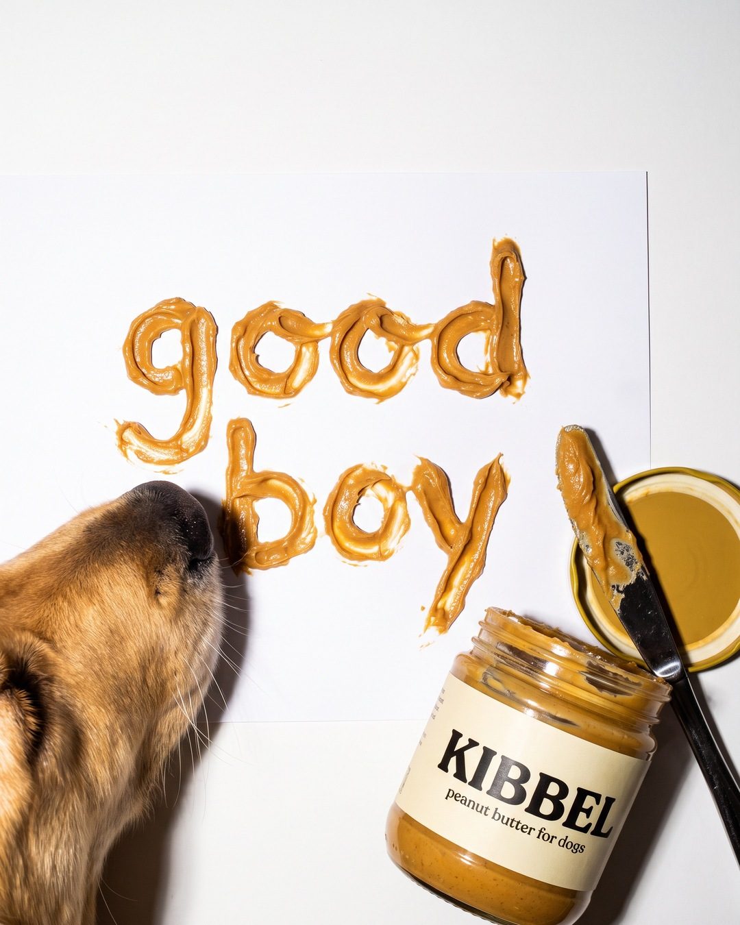 The words good boy piped in peanut butter on white paper beside a KIBBEL jar, a golden retriever sniffing in