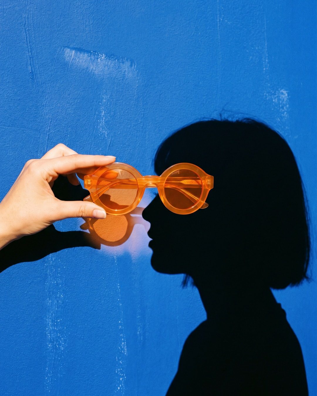 A hand holding orange round sunglasses over the eye of a black profile shadow on a cobalt-blue wall