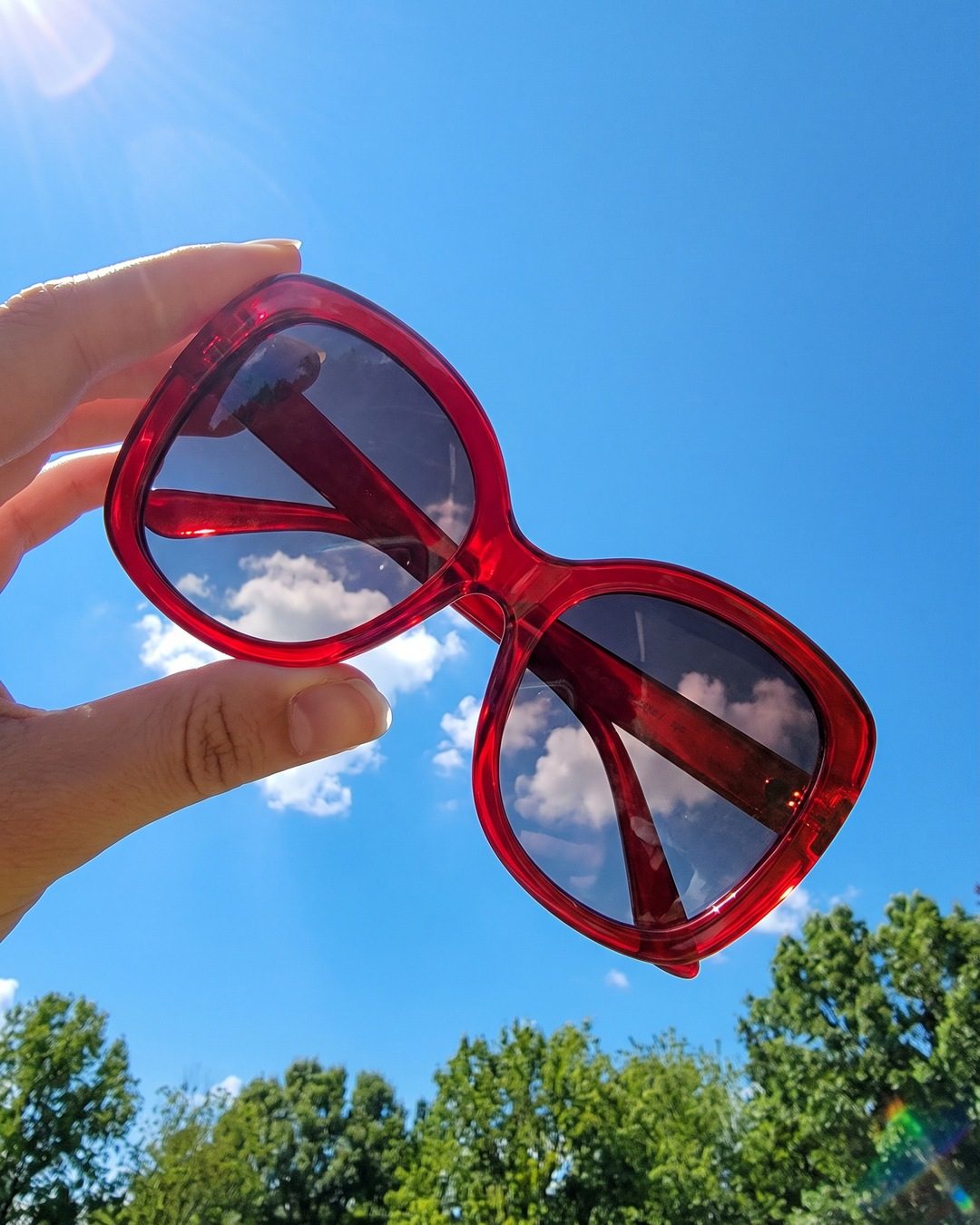 A hand holding red oversized sunglasses up against a blue sky, with clouds reflected in the lenses