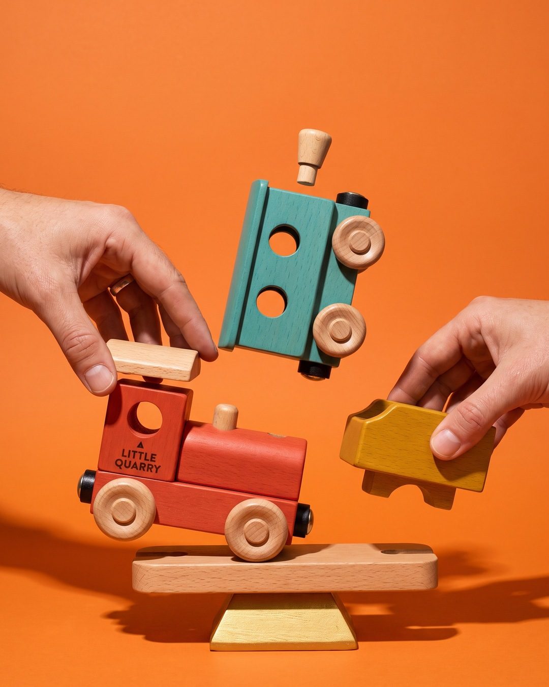 Two hands assembling a red, teal and mustard wooden toy train in mid-air against an orange backdrop