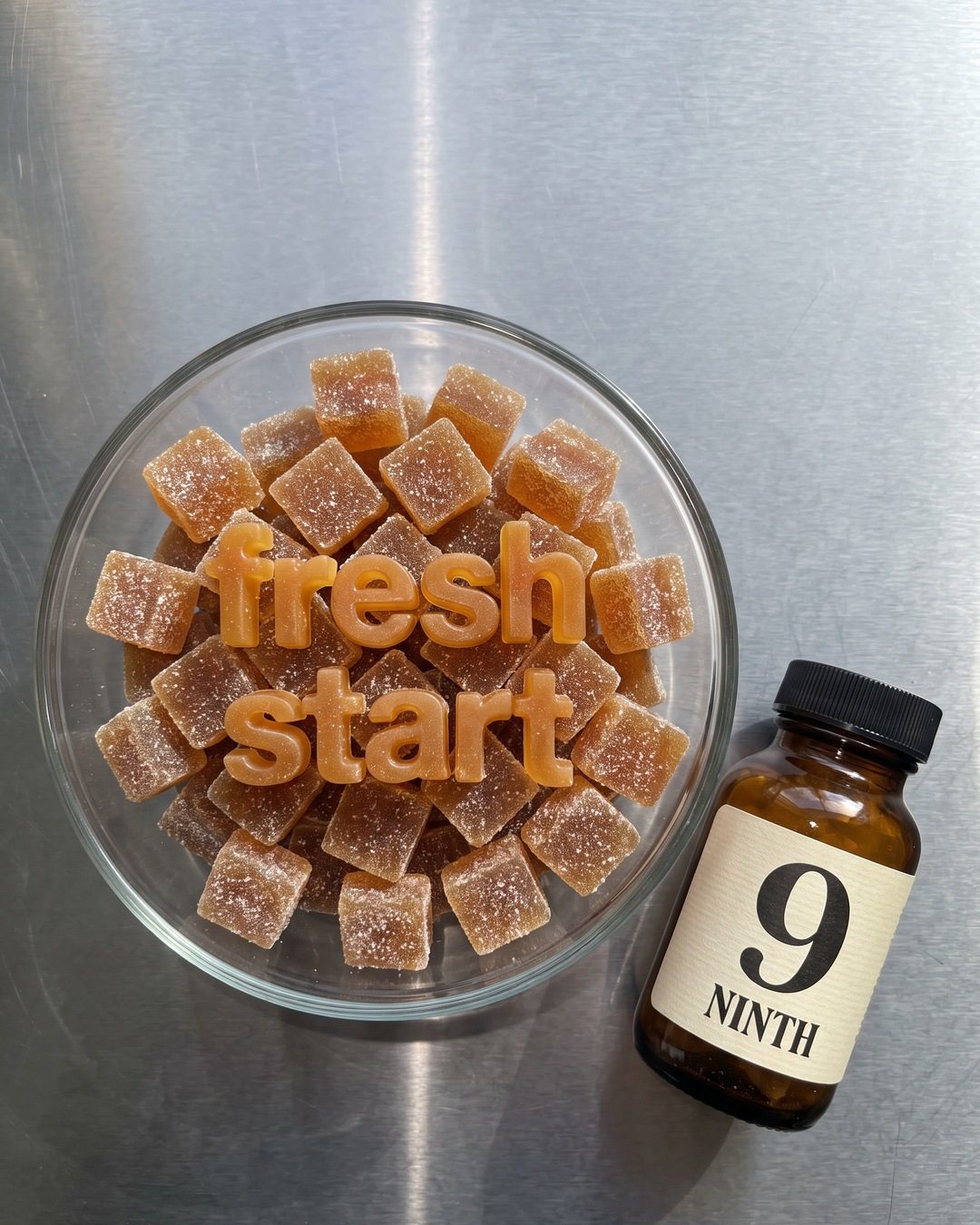 A glass bowl of amber gummies with letter-shaped gummies spelling fresh start, beside a NINTH bottle on steel