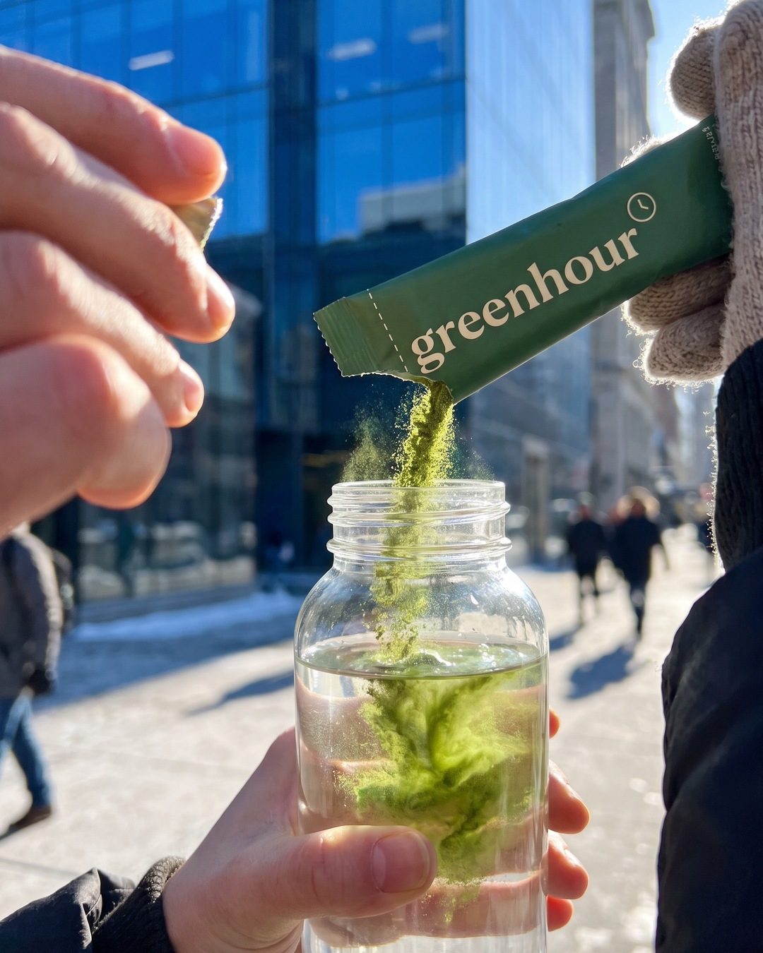 A hand tips a green greenhour sachet so powder pours into a water bottle on a sunny city street
