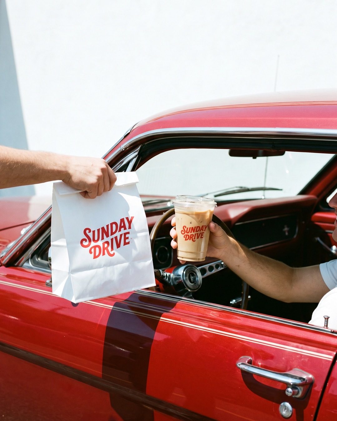 A takeaway bag and iced coffee handed through the window of a red vintage car