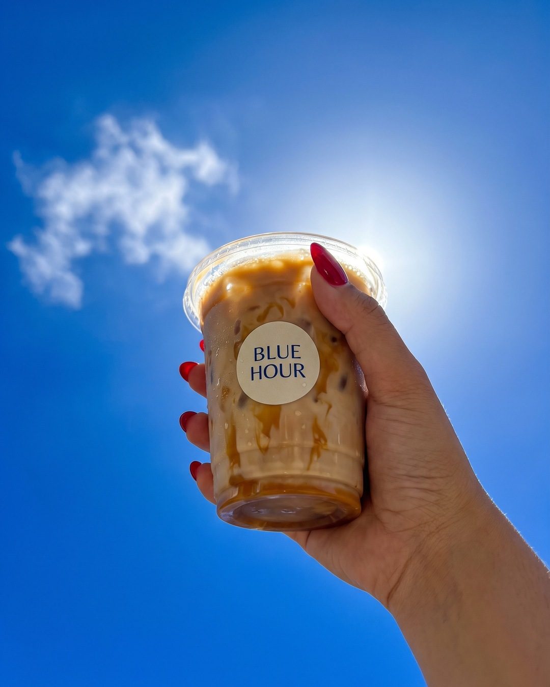 A hand with red nails holding an iced latte up against a bright blue sky with one cloud