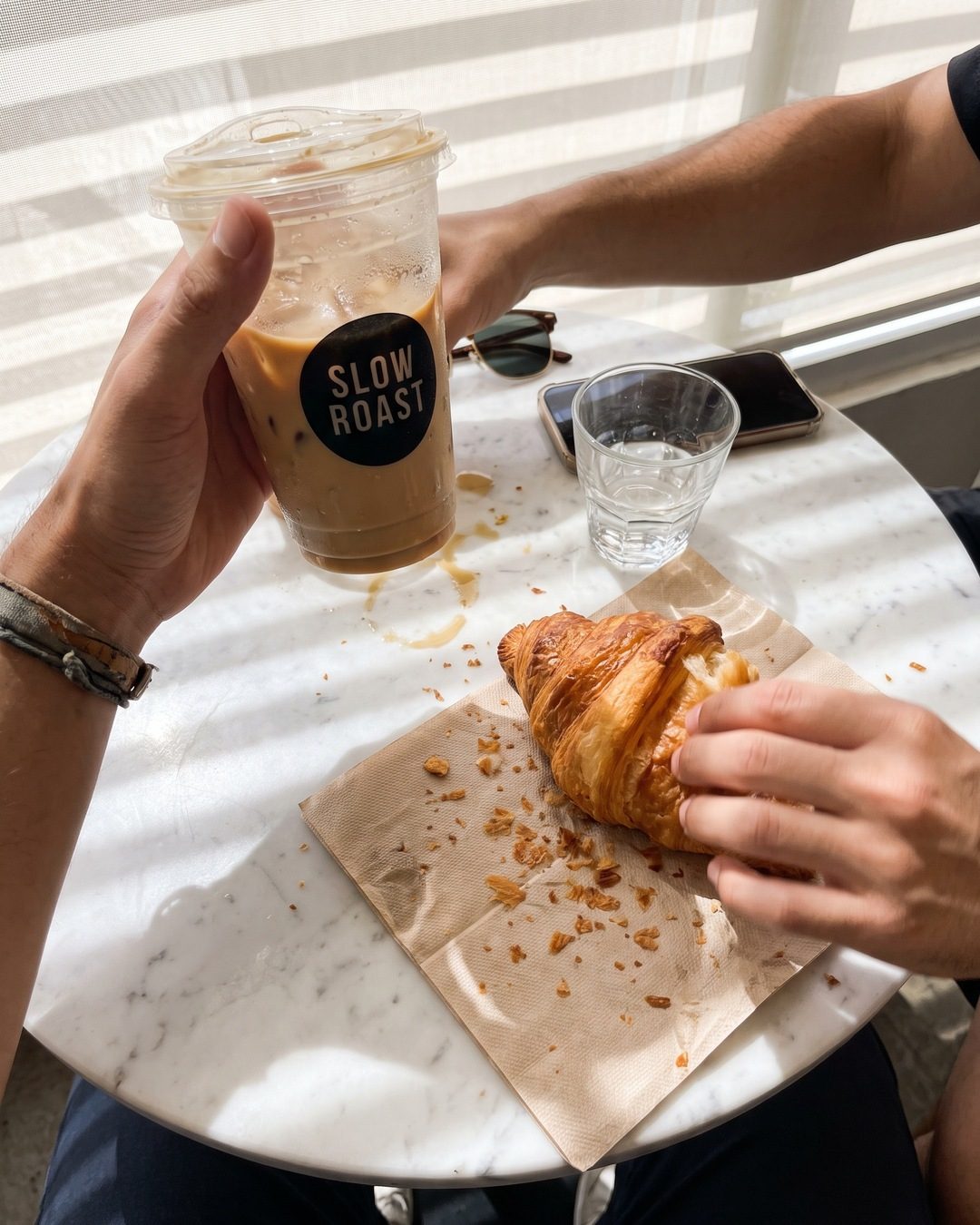 Over-the-table view of a hand holding an iced latte while another tears a croissant on a marble cafe table