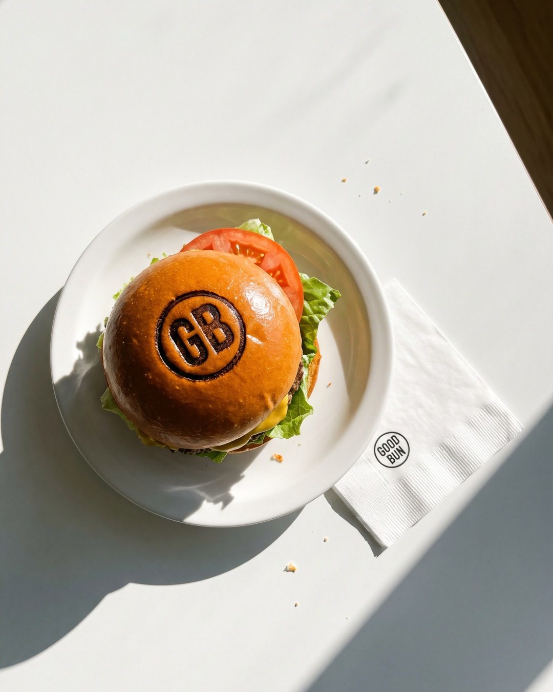 A cheeseburger on a white plate with a GB monogram branded into the bun, next to a GOOD BUN napkin