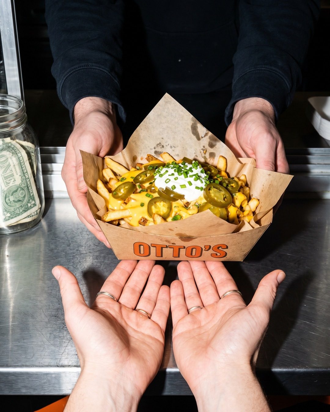 A worker hands a paper boat of loaded fries printed OTTO'S across a steel counter to open palms