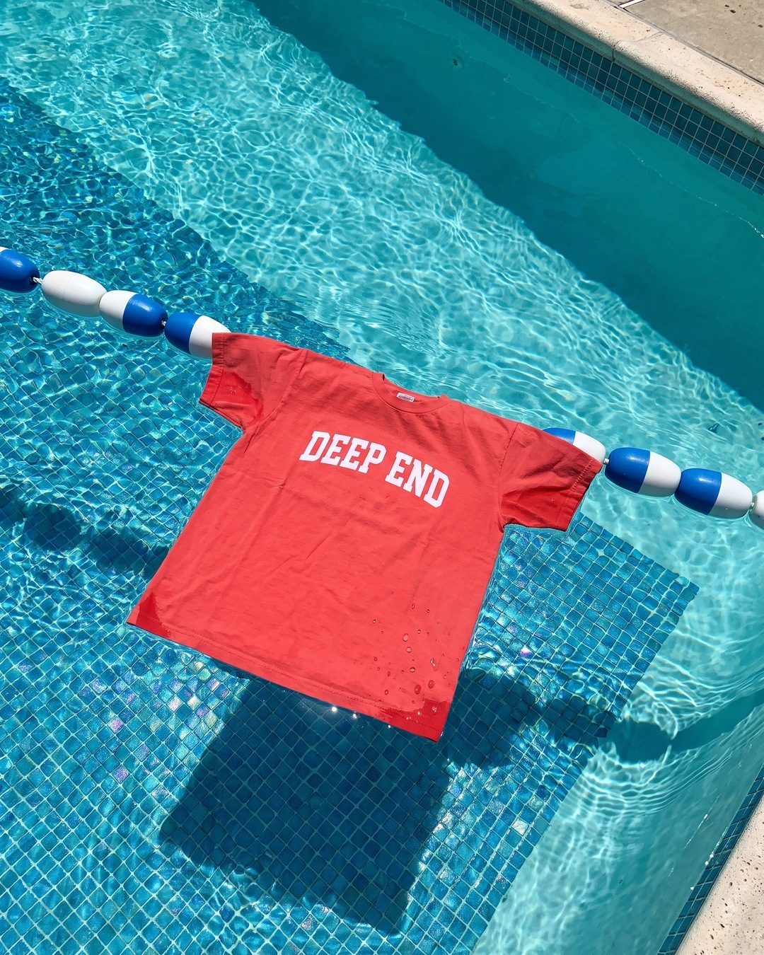 A red DEEP END t-shirt floating on a pool lane rope over blue tiles