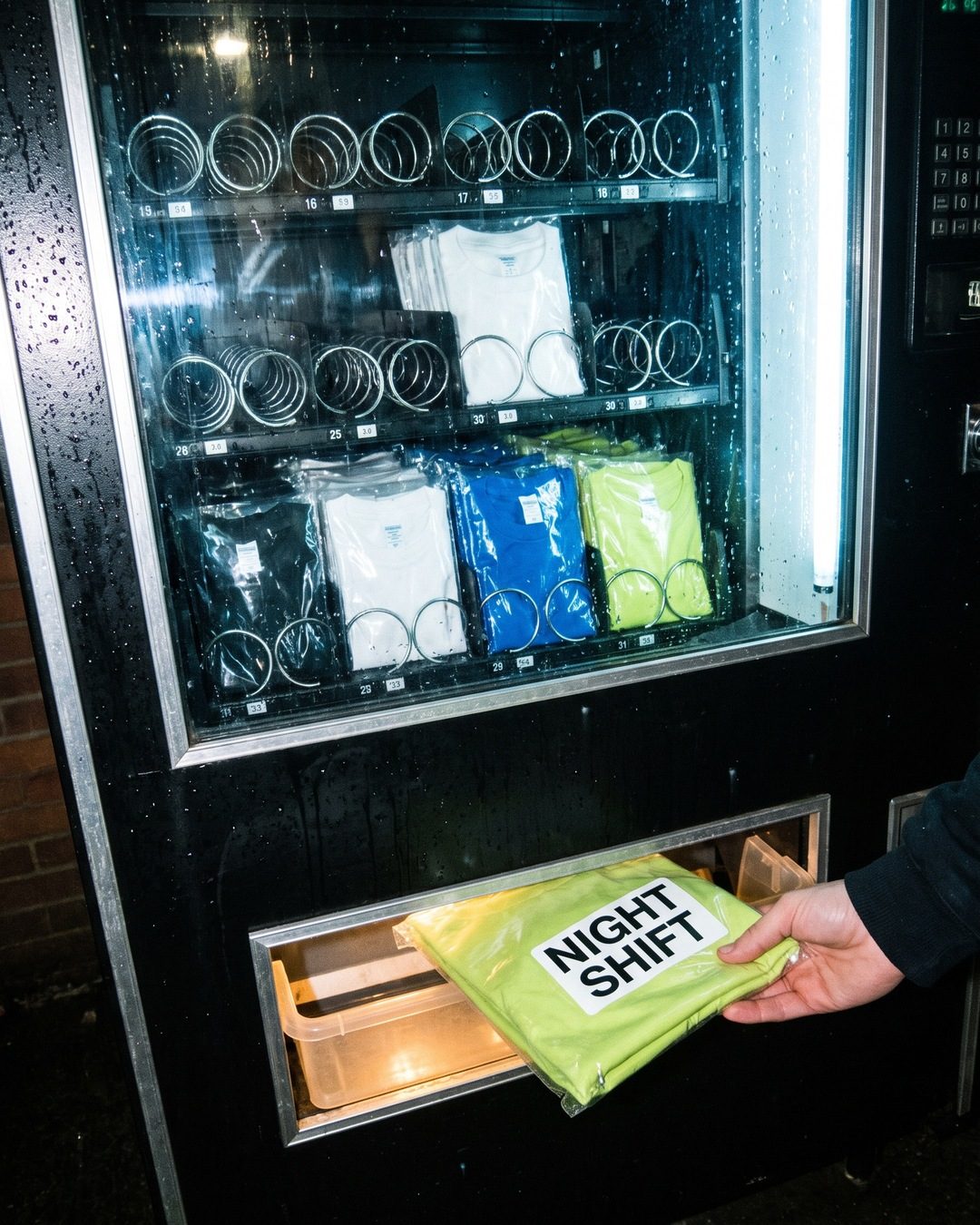 A hand taking a bagged green NIGHT SHIFT t-shirt from the tray of a vending machine stocked with tees