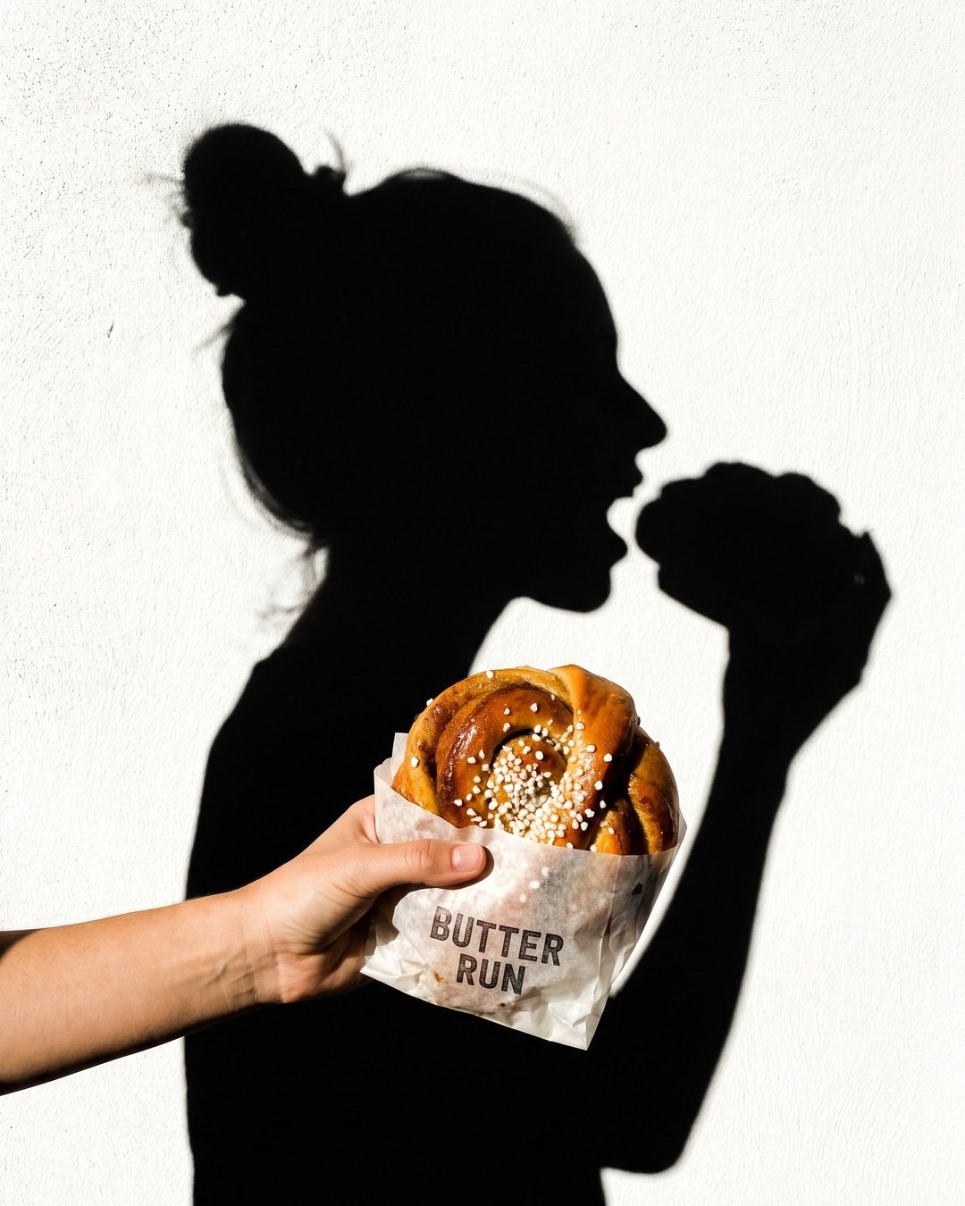 A hand holds a sugared bun in front of a white wall where a woman's shadow is about to bite it