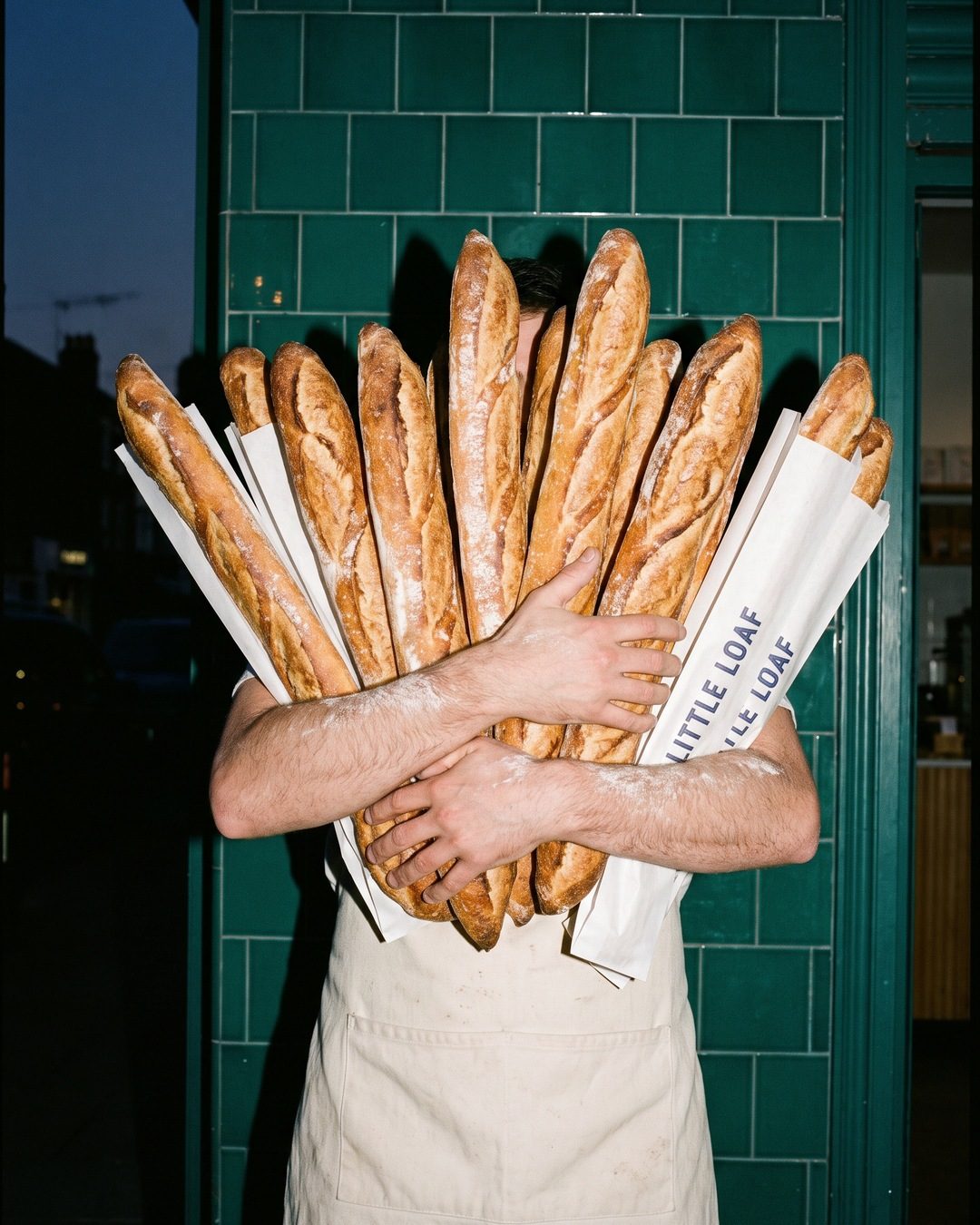 A baker in an apron hugs an armful of baguettes that hides her face, outside a green tiled shop