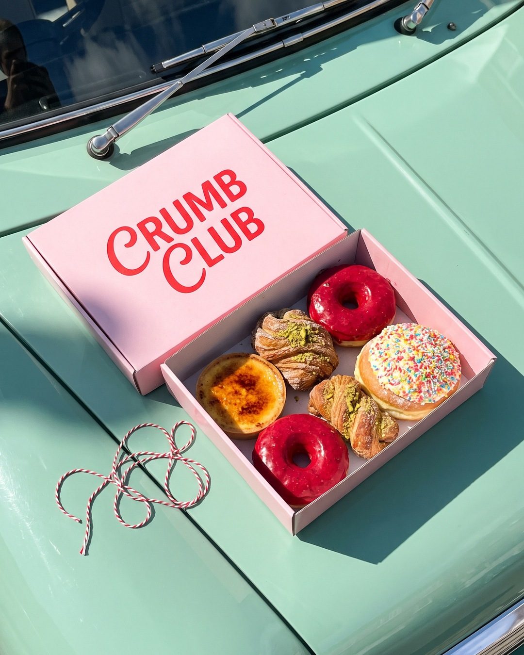 An open pink Crumb Club pastry box of doughnuts and tarts on the bonnet of a mint green car