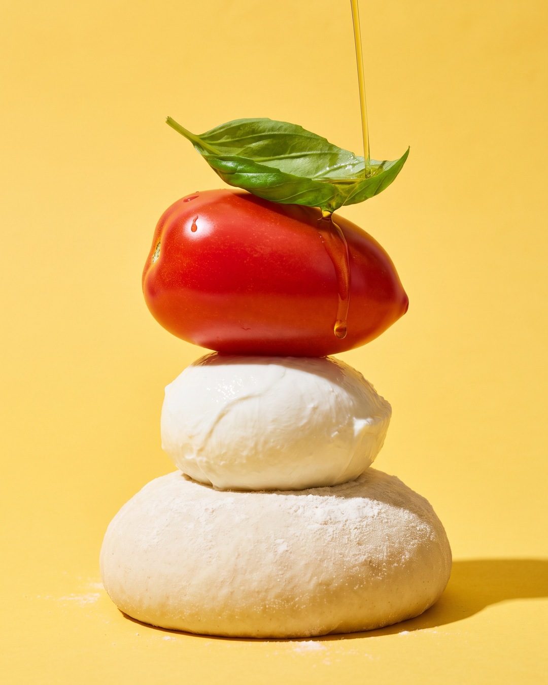 A tower of dough, mozzarella, a plum tomato and basil on a butter-yellow backdrop, olive oil pouring onto it