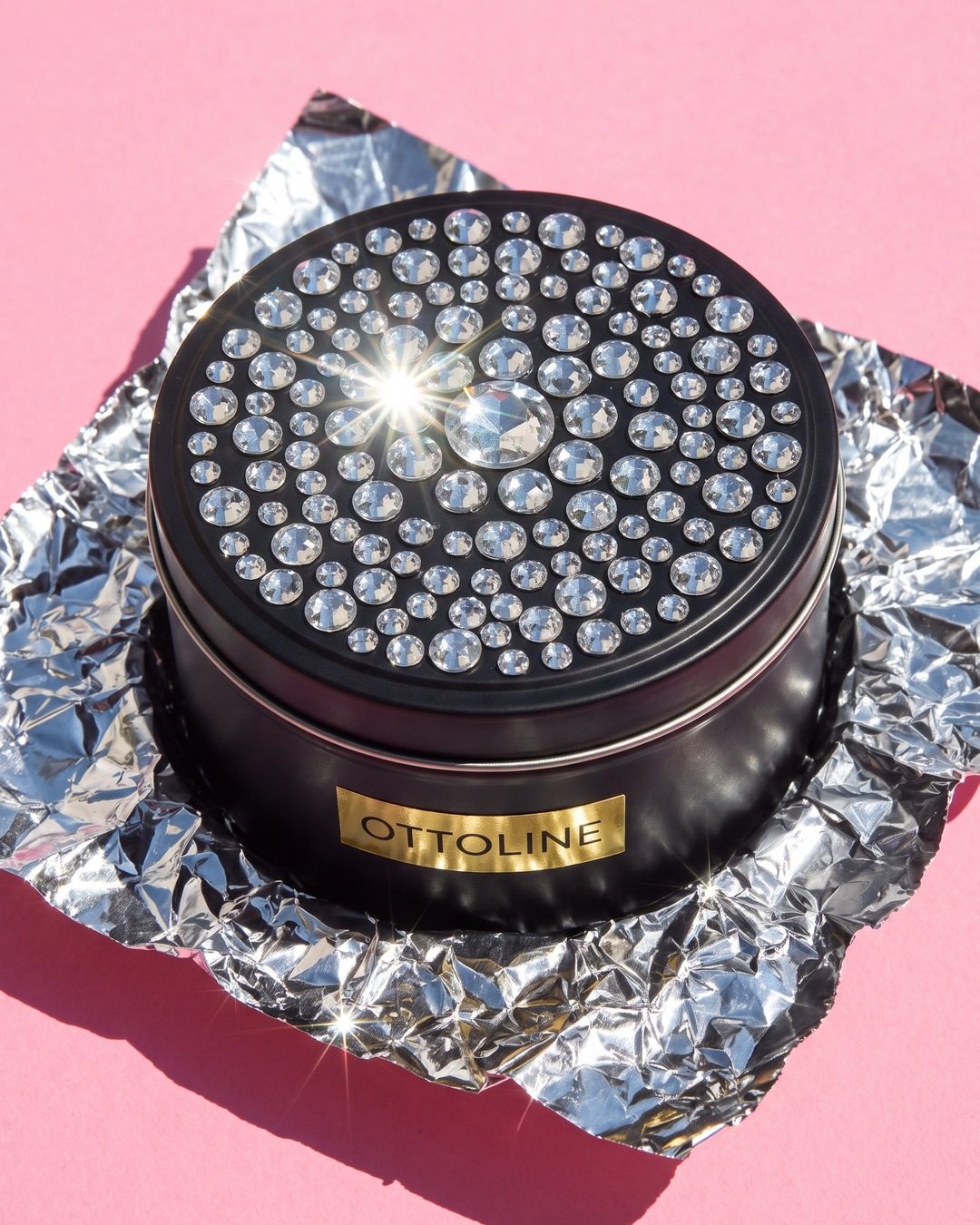 A black candle tin with a rhinestone-covered lid on crumpled foil against hot pink