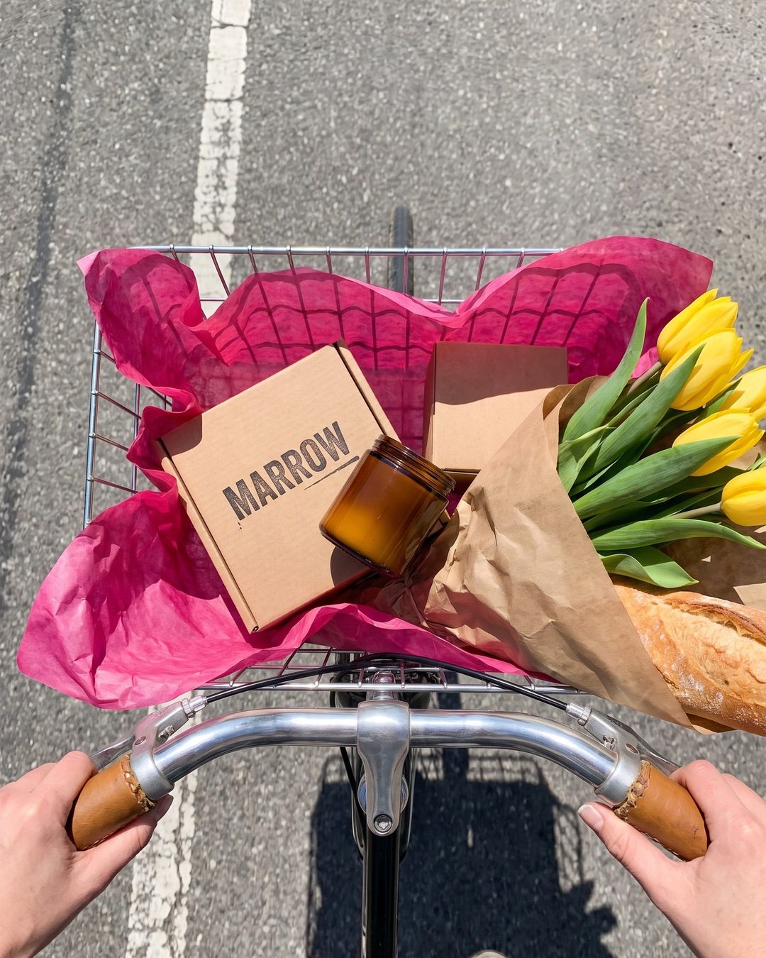 Looking down over bike handlebars at a basket with a Marrow candle box, tulips and a baguette