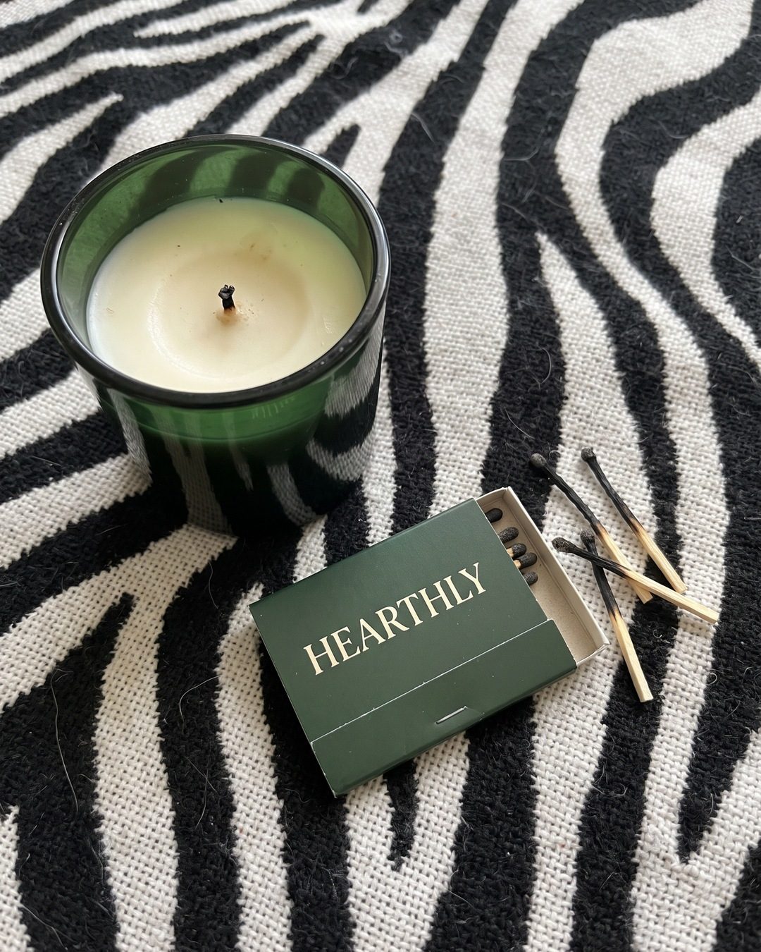 A green glass candle and a matching green Hearthly matchbook with burnt matches on a zebra-print blanket