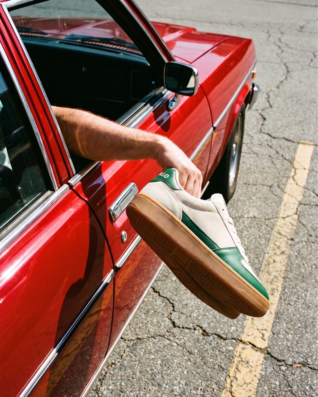 An arm holding a cream and green suede sneaker out of the window of a red vintage car