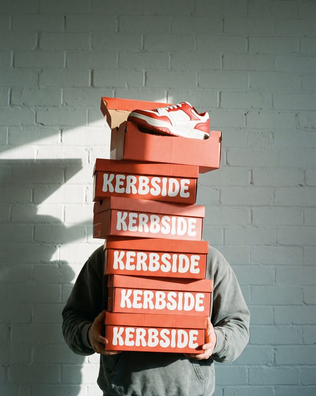 A person's face hidden behind a stack of red KERBSIDE shoeboxes against a white brick wall
