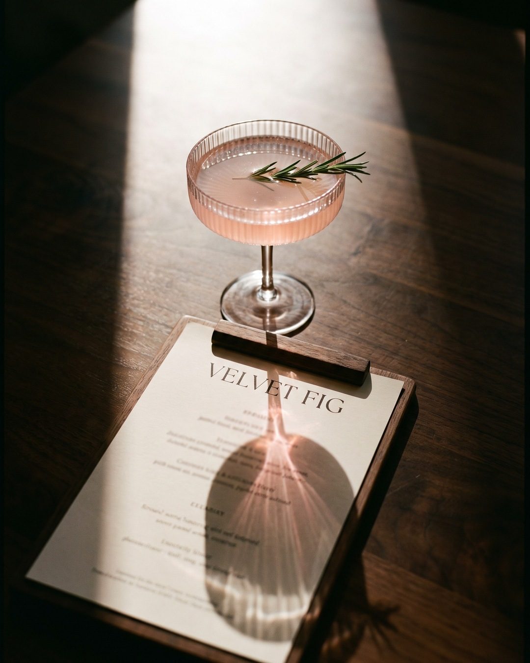A pink coupe with a rosemary sprig casting a rose-gold shadow across a Velvet Fig menu in late sun