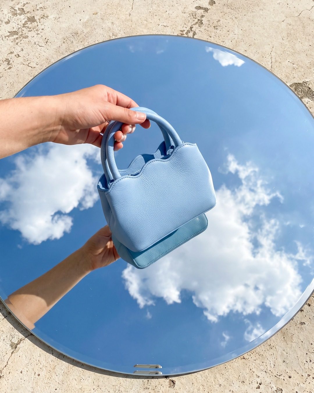 A hand holding a baby-blue scalloped bag over a round mirror on the ground reflecting blue sky