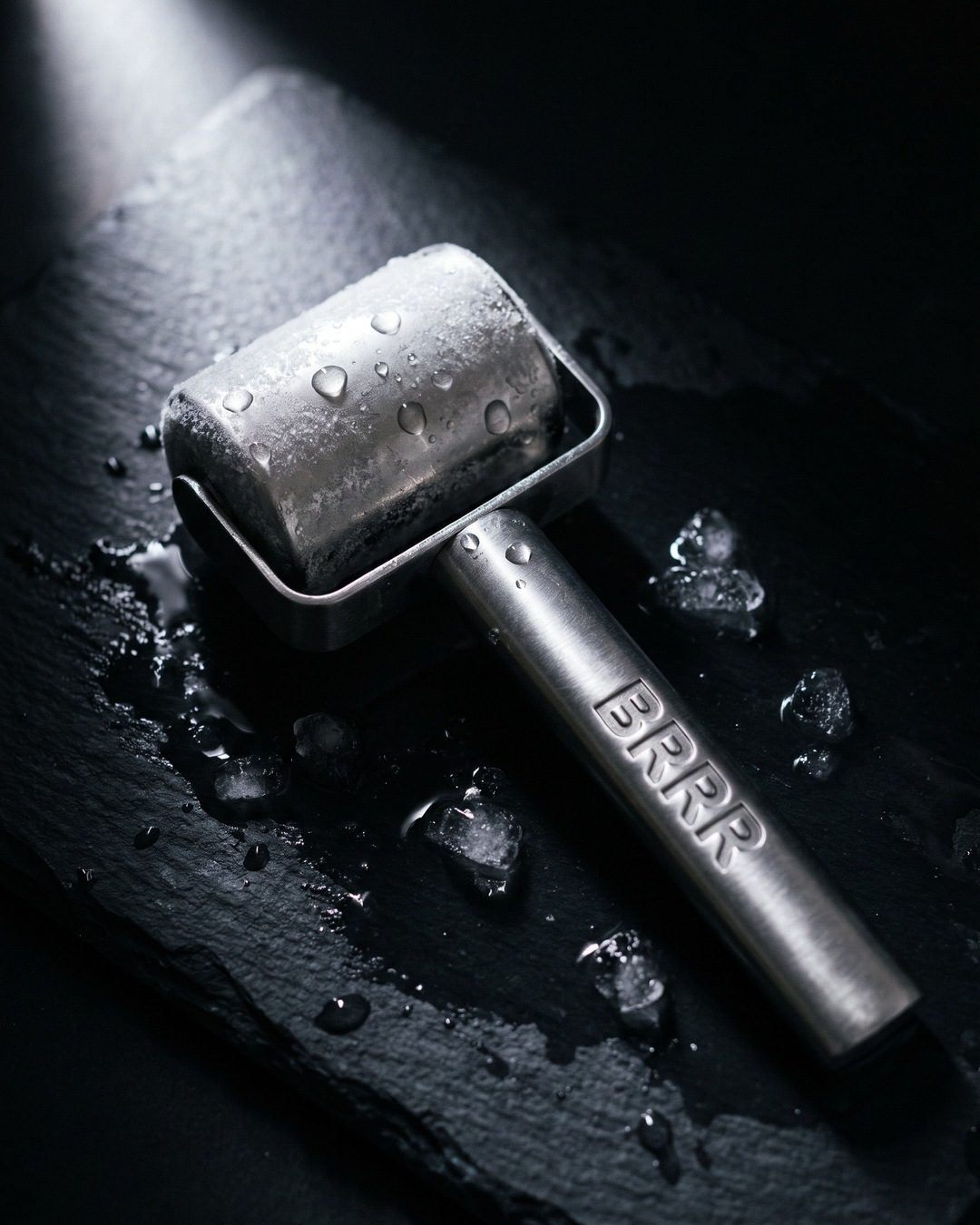 A frosted steel Brrr ice roller covered in droplets on wet black slate among melting ice chips