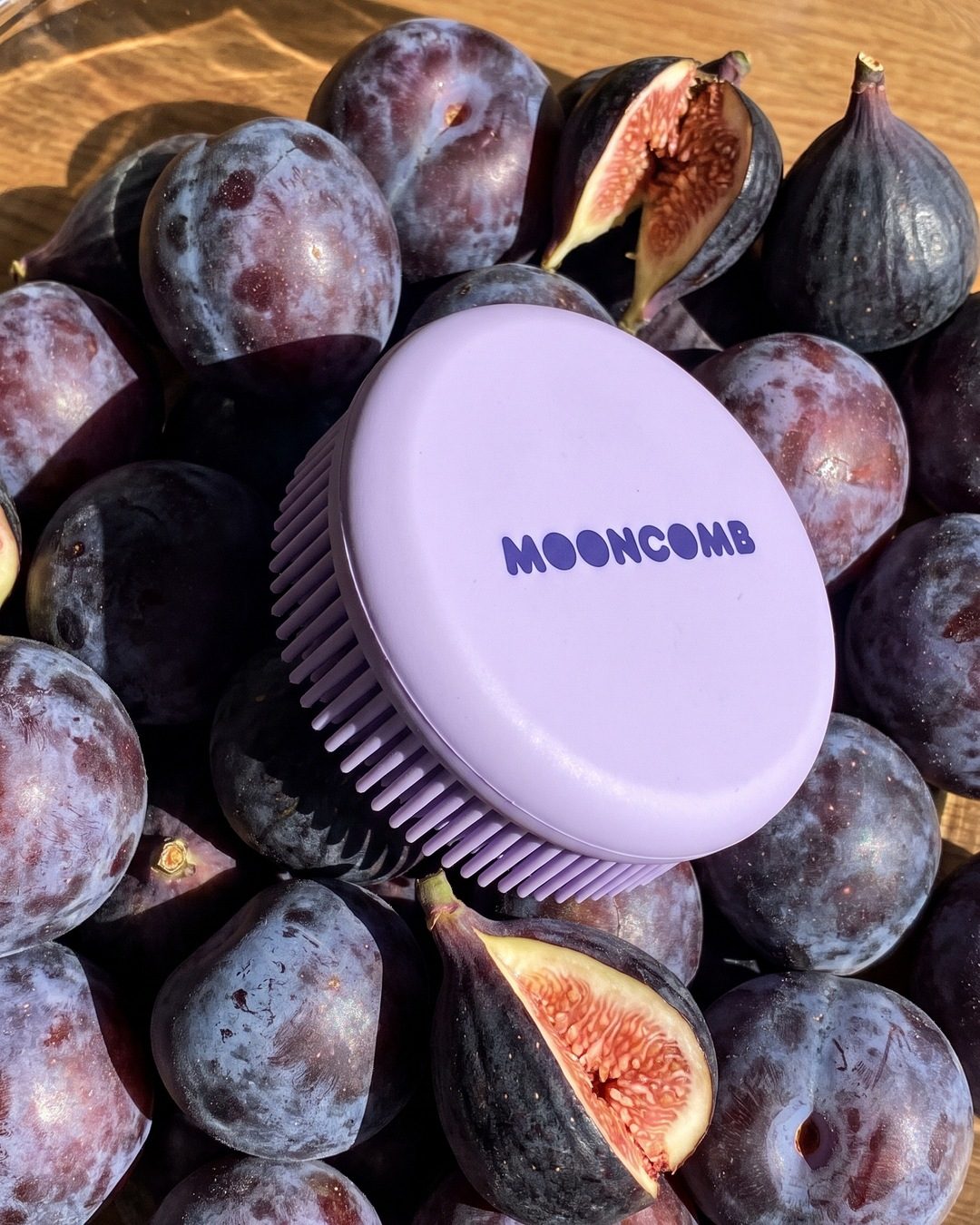 A lavender Mooncomb scalp massager lying on a bed of purple plums and halved figs in hard sun