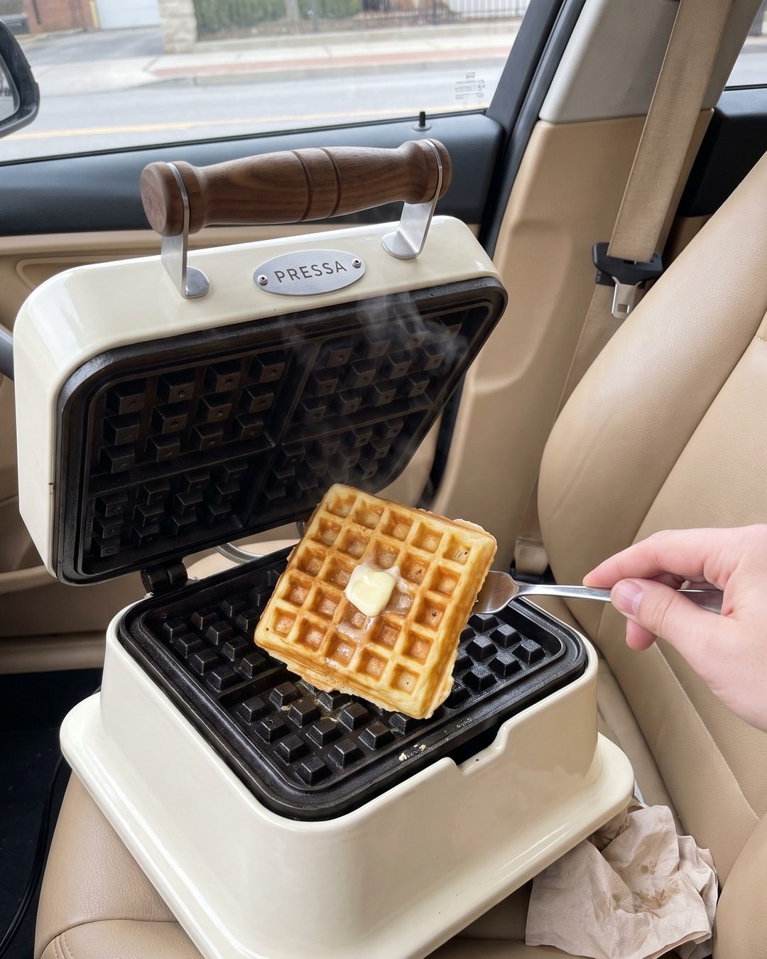 An open cream waffle iron on a car's passenger seat, a hand lifting out a buttered waffle