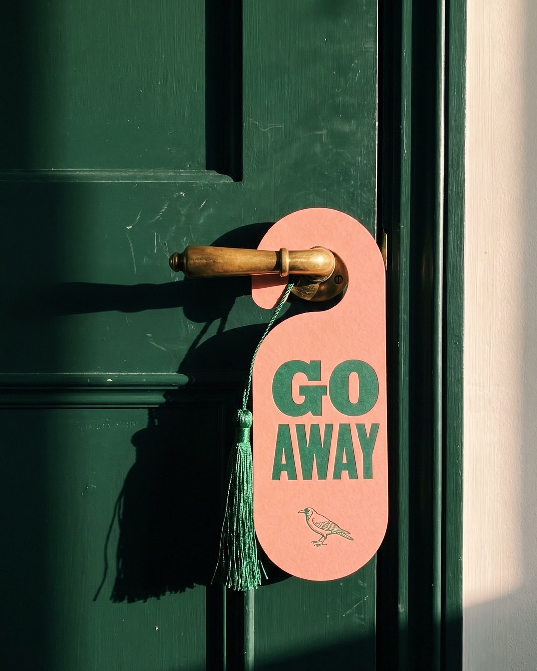 A salmon-pink door hanger reading GO AWAY with a green tassel on a green hotel door in low sun
