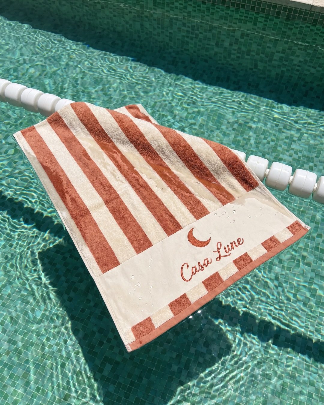 A cream and terracotta striped towel embroidered Casa Lune floating on a pool lane rope