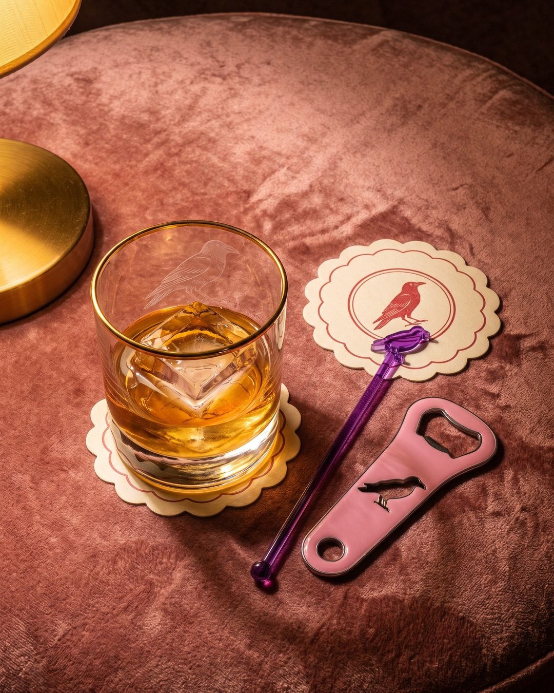 A whisky glass, coasters, a swizzle stick and a pink bottle opener with a crow motif on rose velvet