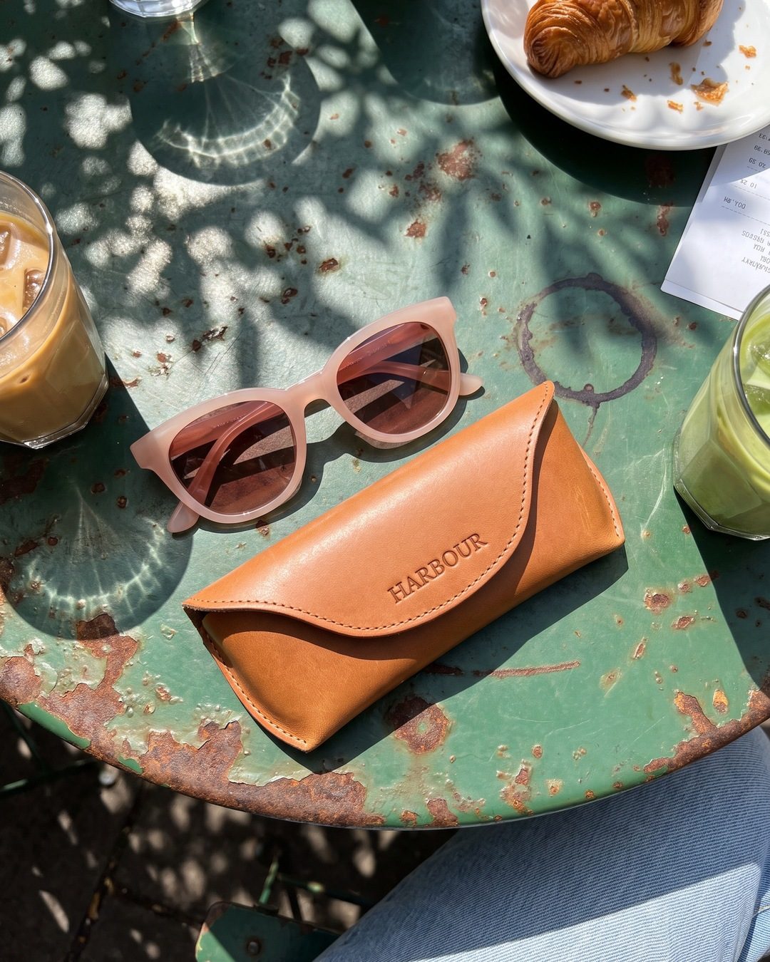 Pink sunglasses and a tan Harbour leather case on a rusty green cafe table with iced drinks and a croissant