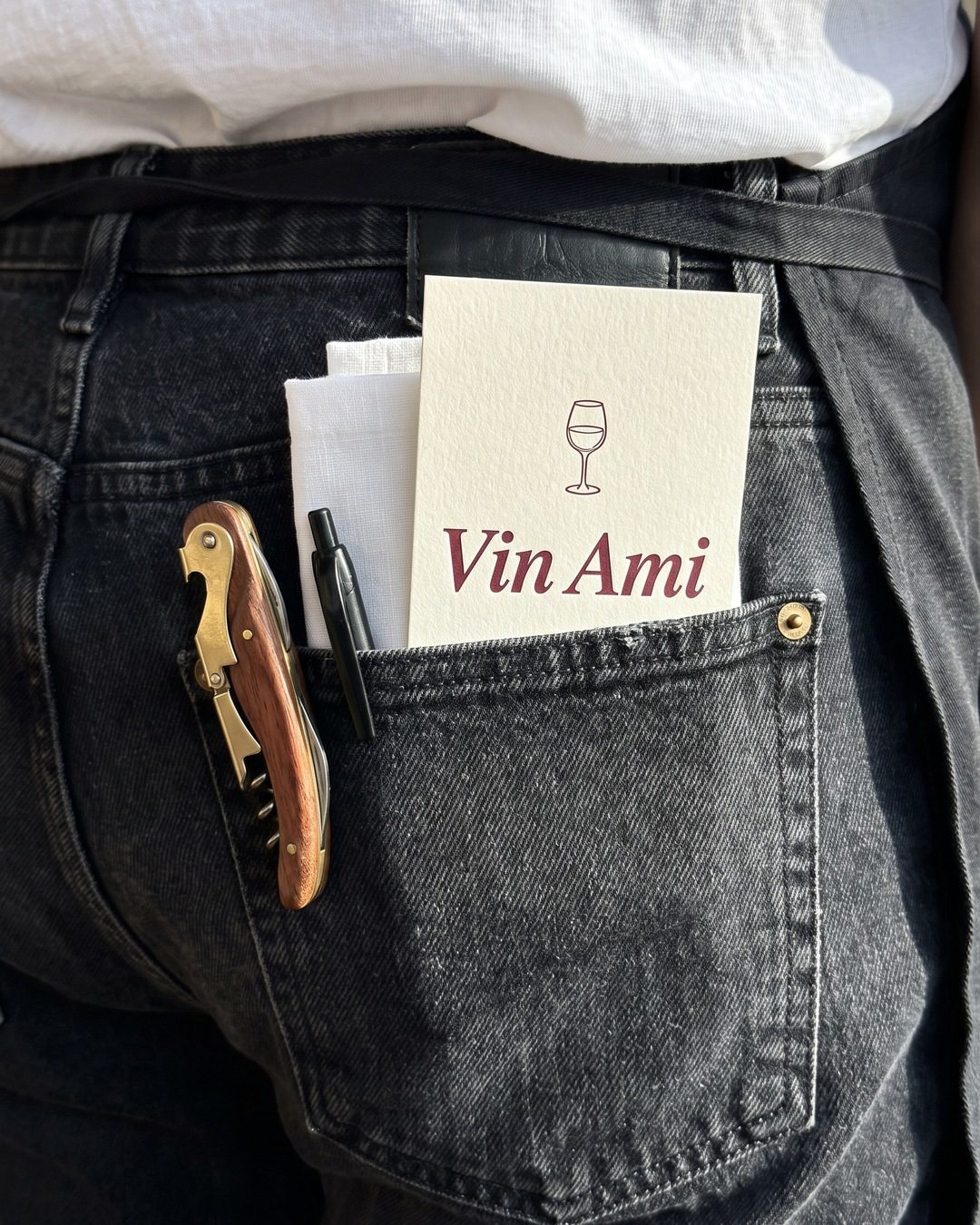 Back pocket of black jeans holding a corkscrew, a pen, a white cloth and a cream Vin Ami card